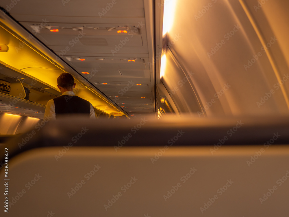 Stewart standing in the aisle of an airplane aircraft cabin. Overhead baggage rack opened and yellow lights turned on. View from behind the passenger seat.