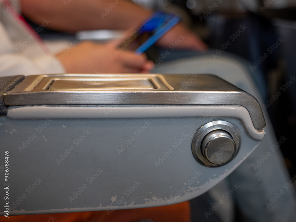 Closeup of a round metal button on an arm support to fall back and have a rest at an airplane. Person sitting in a passenger seat and holding a smartphone.