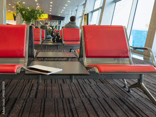 Obraz na płótnie Rows of red seats at an airport terminal. Magazine lying at a table between chairs. People sitting and waiting planes boarding. w salonie