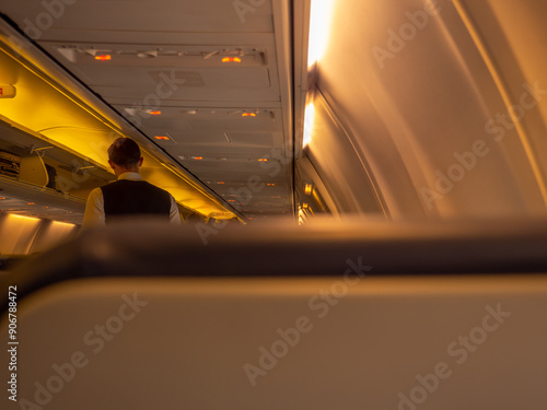 Obraz na płótnie Stewart standing in the aisle of an airplane aircraft cabin. Overhead baggage rack opened and yellow lights turned on. View from behind the passenger seat. w salonie