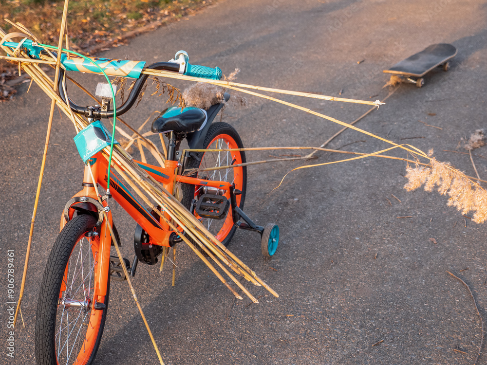 Two-wheeled orange and blue child bike with extra wheels carrying bulrush and pulling a skate board on a rope behind. Child playing active creative games outdoors in a park.