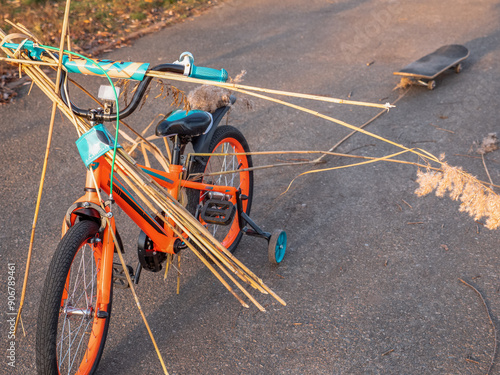 Obraz na płótnie Two-wheeled orange and blue child bike with extra wheels carrying bulrush and pulling a skate board on a rope behind. Child playing active creative games outdoors in a park. w salonie