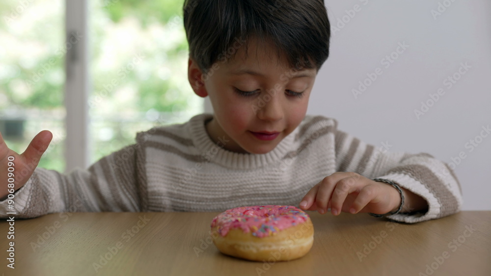 A young child sitting at a table and holding a sprinkled donut, looking at it with curiosity and anticipation, demonstrating delayed gratification, ready to take a bite A young child sitting at a table and holding a sprinkled donut, looking at it with curiosity and anticipation, demonstrating delayed gratification, ready to take a bite
