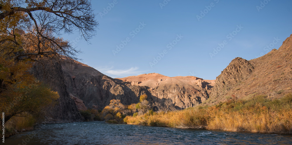 Amber foliage frames a mountains river, meandering at the foot of towering, rugged cliffs under the gentle embrace of a clear azure sky.