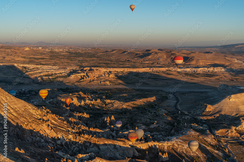 Naklejka premium multiple hot air balloons rise above a stark landscape of eroded rock formations, casting colorful silhouettes against the clear sky.