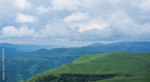 a tranquil panorama, rolling hills and gentle slopes are clad in vibrant green, with distant mountains partially veiled by a soft mist. Above, a dramatic sky, where billowing clouds