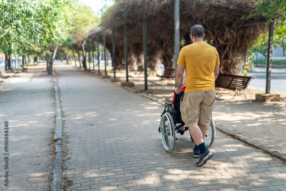 Obraz premium Man on his back pushing a wheelchair with an elderly woman sitting in a public park outside.