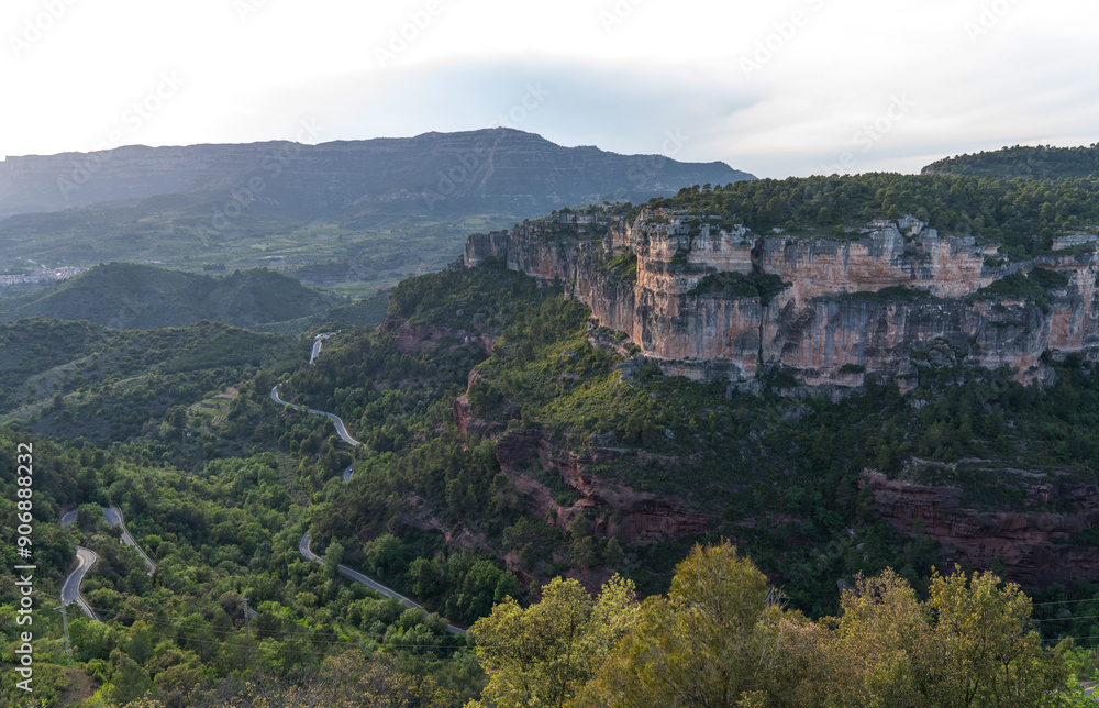 Fototapeta premium breathtaking view of majestic cliffs with layered rock formations, winding roads through lush greenery, and a serene skyline at dusk.