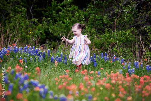 A young girl in a striped sundress smiles, her hands spread out in wonder, stands in a field of orange, red, and blue wildflowers, deep woods at her back.