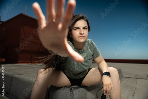 A person wearing a green t-shirt, shorts, and sunglasses confidently poses on a rooftop, reaching out towards the camera against a clear and sunny blue sky. photo