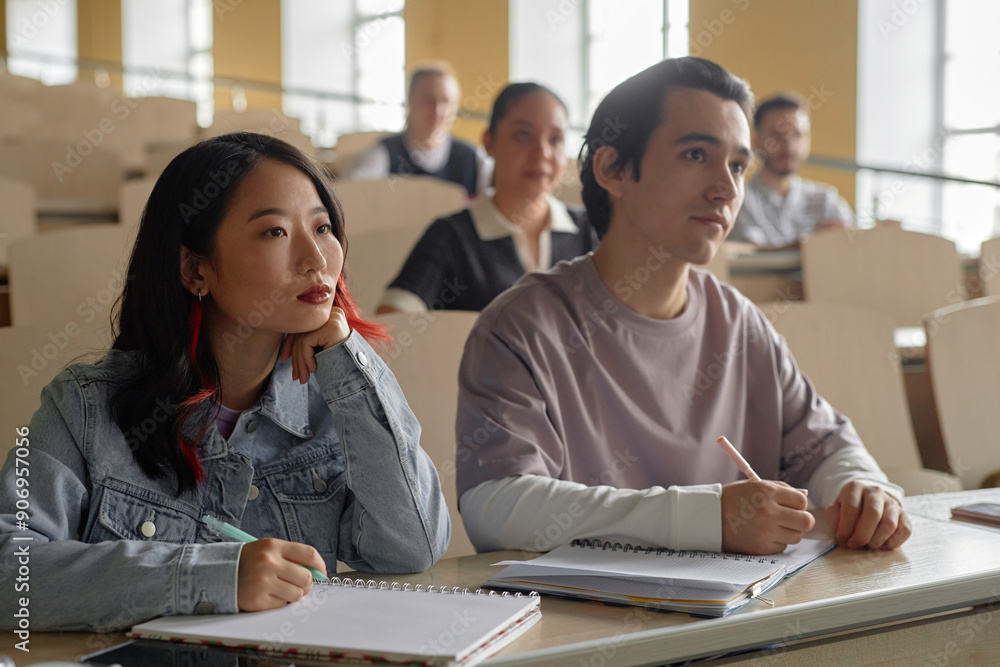 © Seventyfour - Shot of two multiethnic young college students attentively listening to lecture taking notes while sitting at desk in university classroom