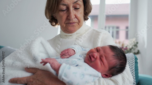 Grandmother trying to comfort crying baby newborn craddling him in her arms standing home living-room. infant during first week of life photo