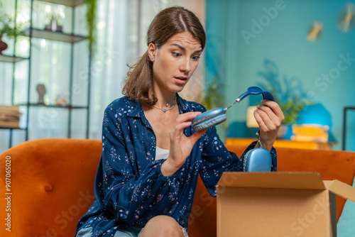 Unhappy young Caucasian woman unpacking carton box, feeling dissatisfied with getting damaged wireless headphones order, writing negative feedback in mobile application, internet shopping concept.
