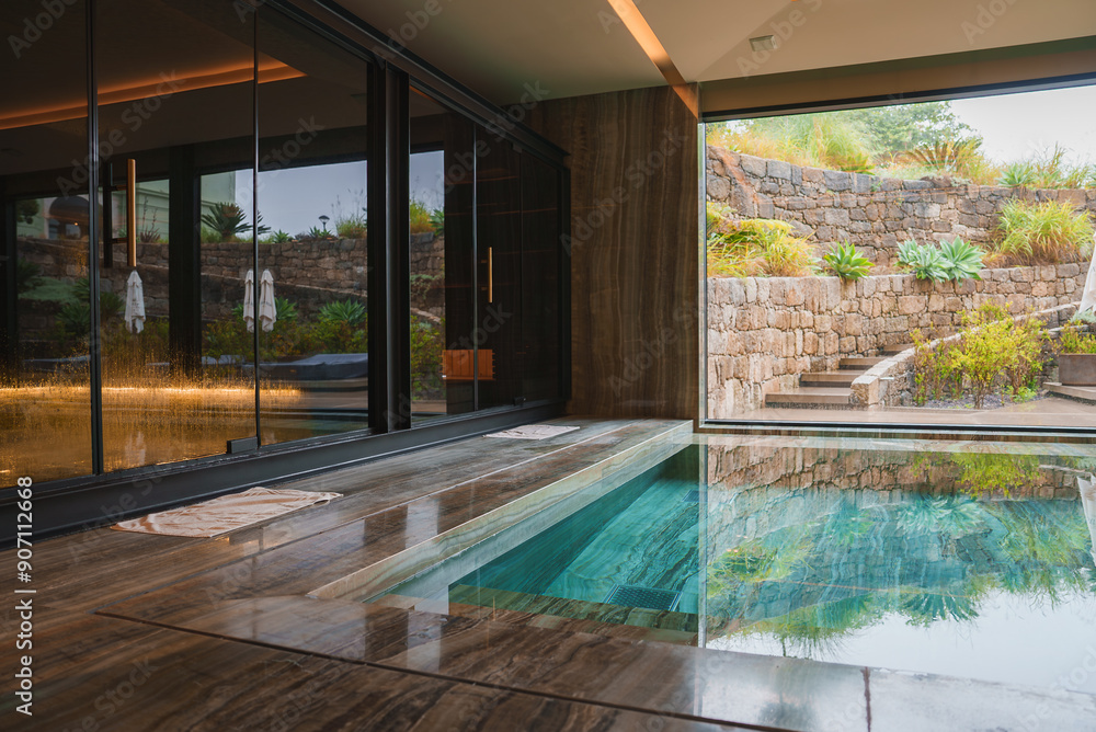 A serene indoor pool area featuring polished stone flooring and walls, large glass doors, and a beautifully landscaped outdoor space with lush greenery.