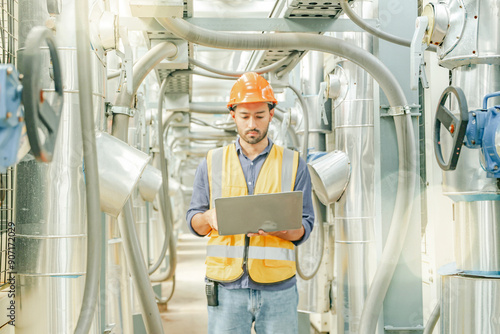 Male HAVC refrigeration system technician stands holding laptop recording preventative inspection refrigeration equipment refrigerant level control valve into the system and checking leaks at various. photo