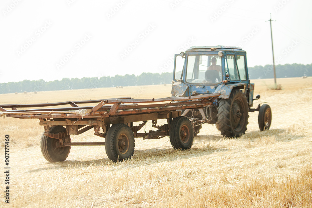 Obraz premium Old Blue Tractor Pulling Wooden Trailer Through Golden Field During Bright Day