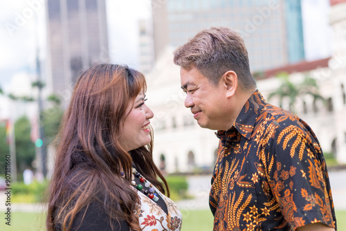 Photography Malay man in his 50s and Chinese woman in her 30s, both dressed in traditional Malaysian attire, gazing into each other's eyes in front of historical building in Kuala Lumpur, Malaysia