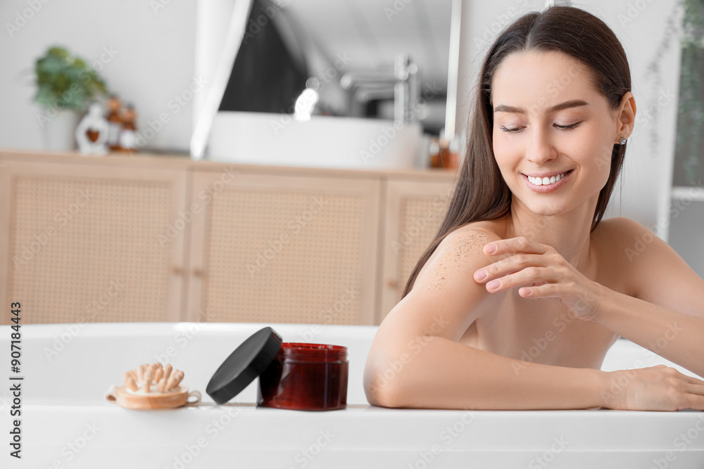 Obraz premium Beautiful young happy woman with jar of body scrub and massage brush sitting in bathroom