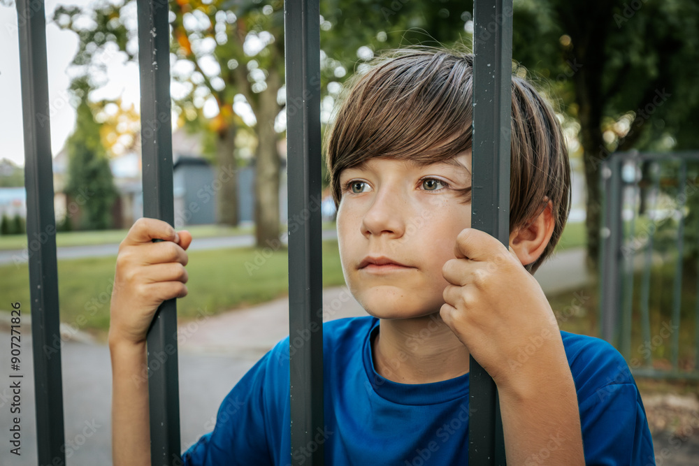 Obraz premium A young boy in a blue shirt grips metal bars while standing in a park, looking directly at the camera. The background features green trees and a fence