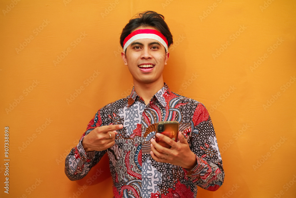Smiling Indonesian men in batik shirts celebrate Indonesian Independence Day on August 17, posing holding mobile phones and pointing with their fingers isolated on yellow background. Smiling Indonesian men in batik shirts celebrate Indonesian Independence Day on August 17, posing holding mobile phones and pointing with their fingers isolated on yellow background.