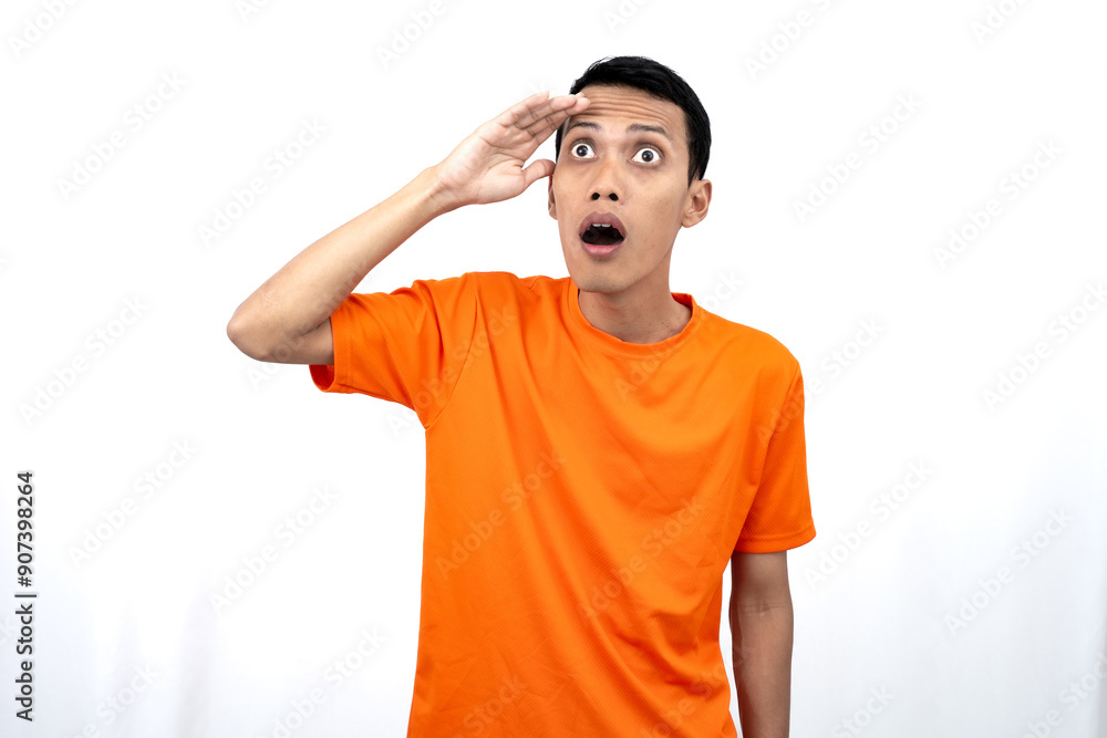 Portrait of a young Asian man wearing an orange t-shirt looking surprised, shocked and shocked. Hands are near eyes, isolated on white background. Portrait of a young Asian man wearing an orange t-shirt looking surprised, shocked and shocked. Hands are near eyes, isolated on white background.