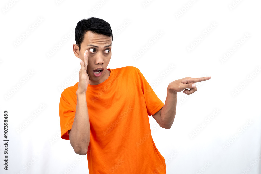 Portrait of a young Asian man wearing an orange t-shirt with his hand near his mouth in a whispering pose while pointing to the side of the copy space. Isolated on white background. Portrait of a young Asian man wearing an orange t-shirt with his hand near his mouth in a whispering pose while pointing to the side of the copy space. Isolated on white background.