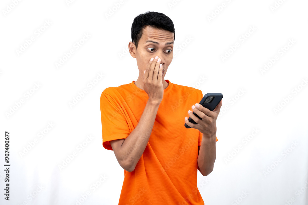 Portrait of a young Asian man wearing an orange t-shirt with a happy expression, surprised to get happy news via smartphone. Isolated on white background. Portrait of a young Asian man wearing an orange t-shirt with a happy expression, surprised to get happy news via smartphone. Isolated on white background.