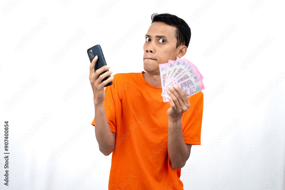 Portrait of a young Asian man wearing an orange t-shirt with a happy, surprised, pleased expression while carrying a smartphone and hundreds of thousands of rupiah. isolated on a white background. Portrait of a young Asian man wearing an orange t-shirt with a happy, surprised, pleased expression while carrying a smartphone and hundreds of thousands of rupiah. isolated on a white background.