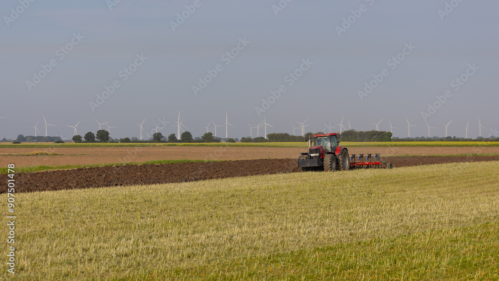 Obraz premium a red tractor is in the process oh plowing. In the distance are wind turbines. A natural setting with copy space in the picture