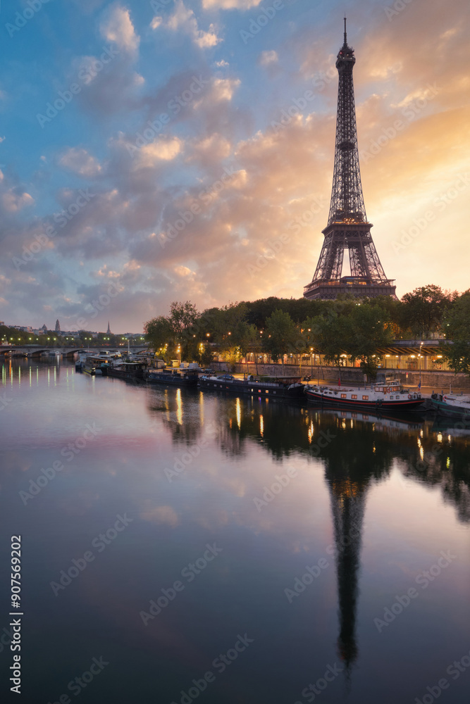 Naklejka premium Eiffel Tower at golden hour with vibrant clouds and its reflection in the Seine River, Paris. The calm water and the lights create a serene and picturesque scene.