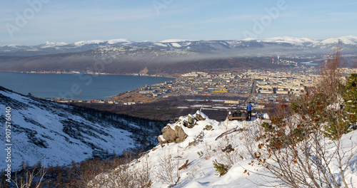 View from the mountain to the city of Magadan. Tourists admire the city from the snow-covered slope of the hill. Young men sit on a bench and look into the distance. Magadan region, Far East of Russia photo