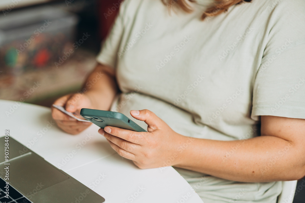 Obraz premium Cropped unrecognizable woman doing online shopping over laptop with credit card and phone at desk in home office. One woman is buying online on her laptop