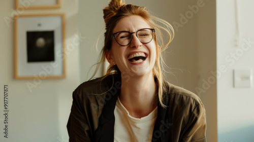 A beaming woman with glasses and a top bun laughs heartily, bathed in warm sunlight in a casual and cozy interior setting.