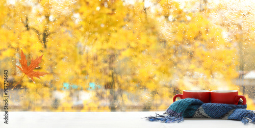 pair of red mugs wrapped in blue scarf against window after autumn rain. warming coffee with maple syrup