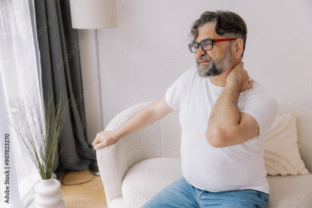 A senior man is sitting indoors, visibly suffering from neck pain. He is holding his neck with a pained expression, indicating discomfort. A senior man is sitting indoors, visibly suffering from neck pain. He is holding his neck with a pained expression, indicating discomfort.