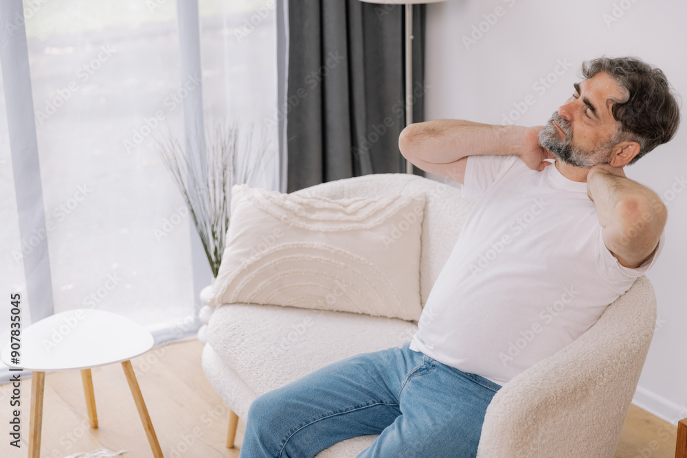 A senior man is sitting indoors, visibly suffering from neck pain. He is holding his neck with a pained expression, indicating discomfort, copy space for your advertising A senior man is sitting indoors, visibly suffering from neck pain. He is holding his neck with a pained expression, indicating discomfort, copy space for your advertising