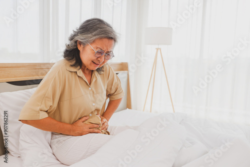фотографія Elderly Asian woman had stomach pain with pained expression on her face while sleeping on the bed, holding her stomach with her hands in excruciating pain for no apparent reason
