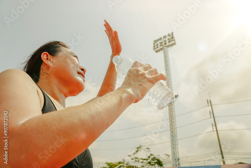 58 year old asian woman exercises under the hot midday sun stand holding bottle of water quench your thirst and refresh your body and use your hands shield your forehead from the sun prevent sunburn. photo