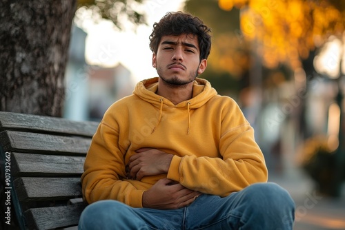 A young Hispanic man is sitting on a bench outside and holding his stomach with his hands. Suffers from pain, discomfort, feels bad, Generative AI