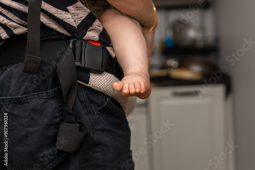 The mother carries the baby in a forward-facing carrier while standing in the kitchen. dangling baby's leg, The image highlights the dual responsibilities of raising children and managing household du