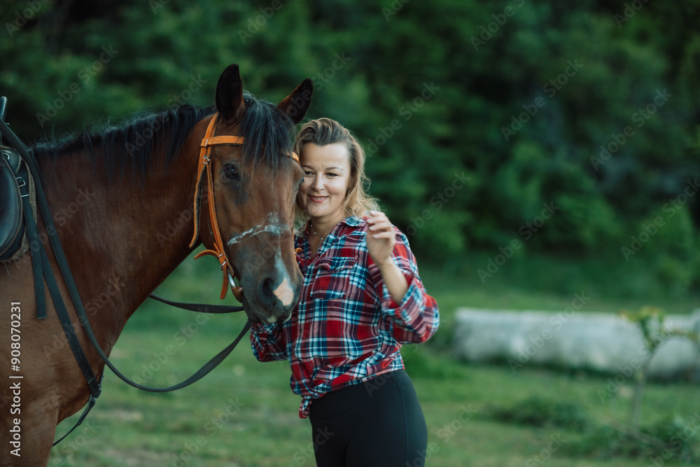 Obraz premium Happy blonde with horse in forest. Woman and a horse walking through the field during the day. Dressed in a plaid shirt and black leggings.