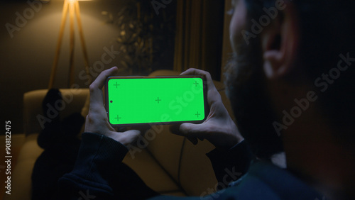Man lying on couch using smartphone horizontally with chroma key green screen. Male watching series or video at home at night.	
