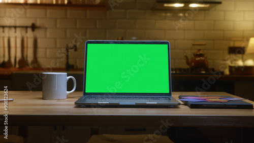 A laptop with a green screen on a kitchen table in kitchen with a coffee mug and notebook.

