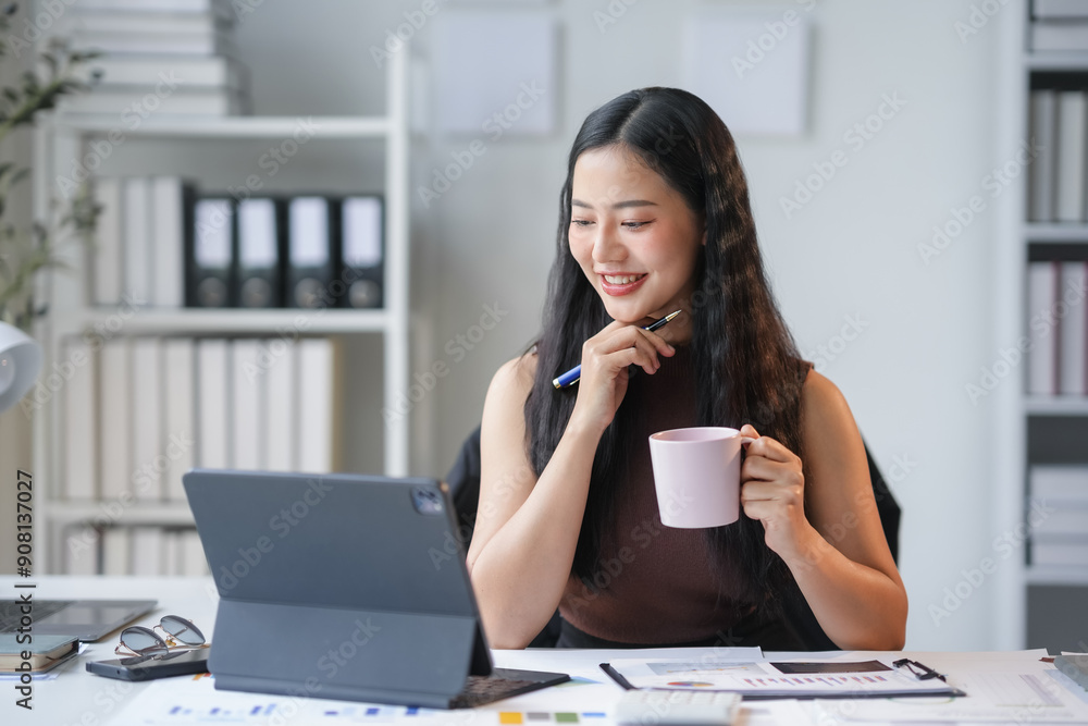 Young businesswoman is smiling while drinking coffee and working from her tablet in a bright and modern office Young businesswoman is smiling while drinking coffee and working from her tablet in a bright and modern office