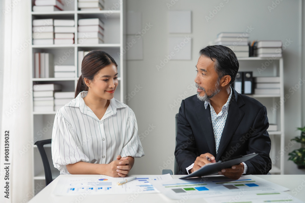 Manager holding a file discussing charts with a young intern while sitting at an office desk Manager holding a file discussing charts with a young intern while sitting at an office desk