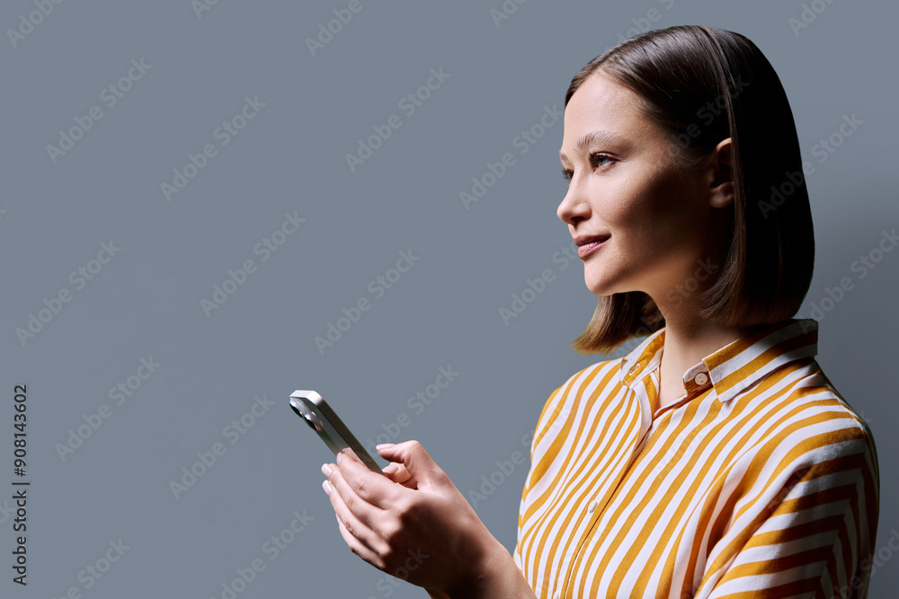 Young confident smiling woman holding smartphone on grey background, profile view Young confident smiling woman holding smartphone on grey background, profile view