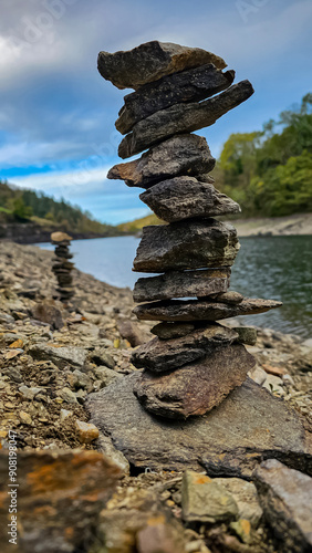 Portrait view of sculpture of towers of stones on shore of beautiful lake; close-up of carefully balanced pile of rugged stones on shore of peaceful river, second tower in background, under blue sky. photo