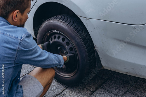 Young man in unscrewing lug nuts on car wheel in process of new tire replacement,using wrench while changing flat tire on the road.