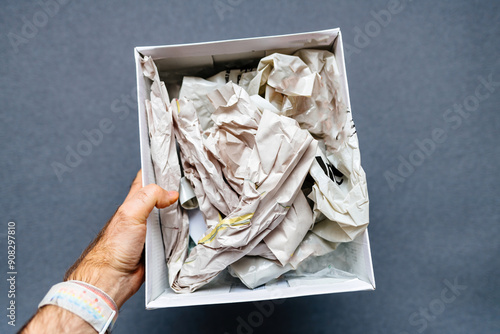 A male hand holding a paper box filled with newspaper against a gray background, in the process of unboxing a fragile object, showcasing careful handling and protection