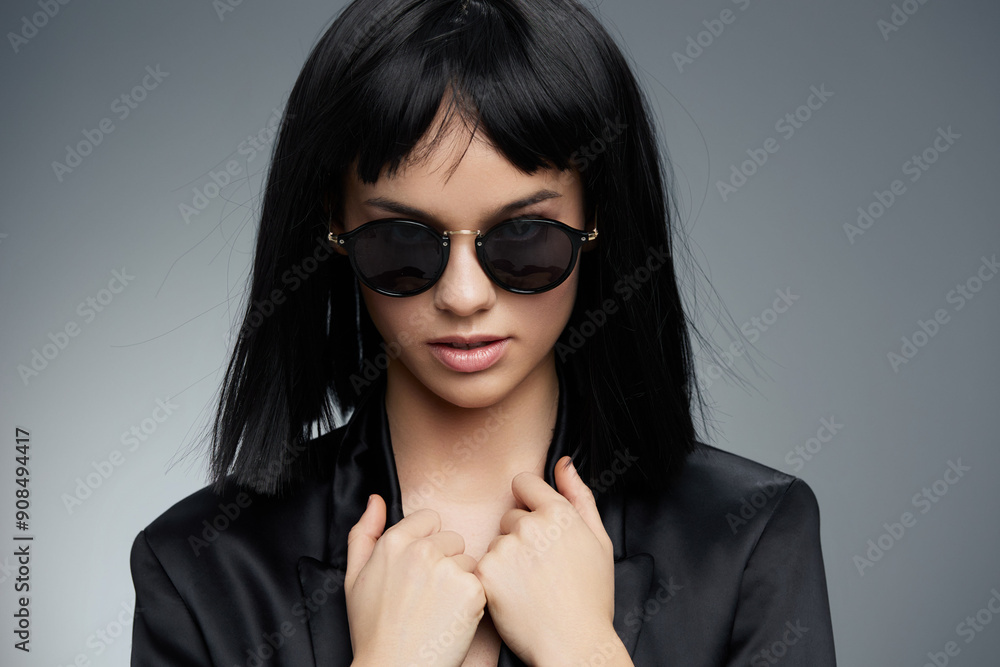 Young woman with black hair, sunglasses, and a black blazer posing confidently against a gray background Young woman with black hair, sunglasses, and a black blazer posing confidently against a gray background
